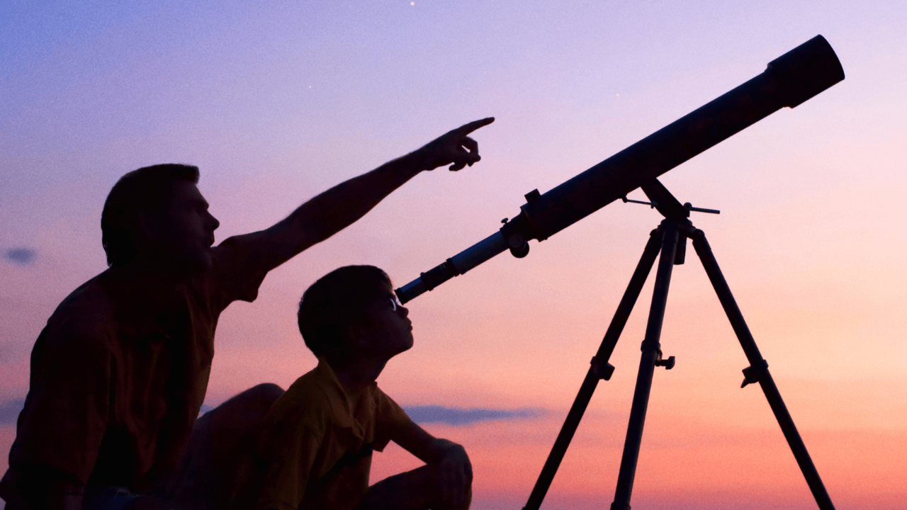 Silhouette of a man and child looking through a telescope at dusk