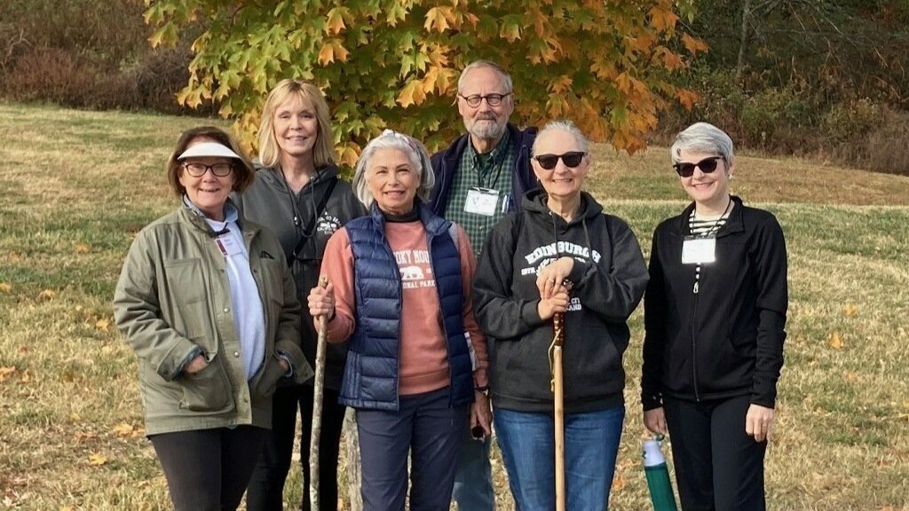 Group of OLLI members wearing casual outdoor clothing and standing together beneath a vibrant autumn tree on grassy park ground