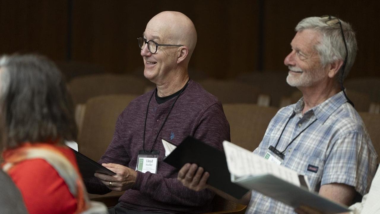Two individuals seated in a classroom setting, smiling and holding folders while wearing name badges.