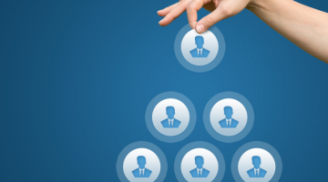 Pictured: On a blue background, a hand is placing an icon of a person at the top of a pyramid. The pyramid represents the hierarchical structure of a workplace.