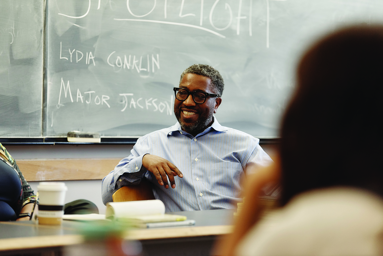 A man sitting in a classroom in front of a chalkboard that reads "Major Jackson"