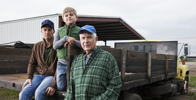 Three generations of men at the back of a truck on the family farm. The grandfather, a senior man in his 60s, is standing with his hand in his pocket, looking at the camera. His adult son and young grandson are behind him on the truck. They are wearing jeans, work boots and jackets or flannel shirt. Everything is brown or green, earthy colors.