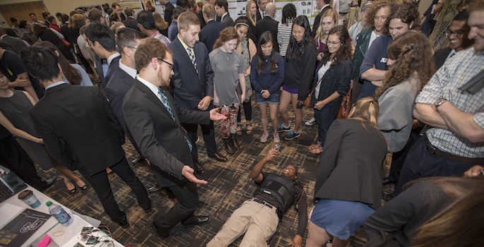 Student in vest lying down while visitors look on