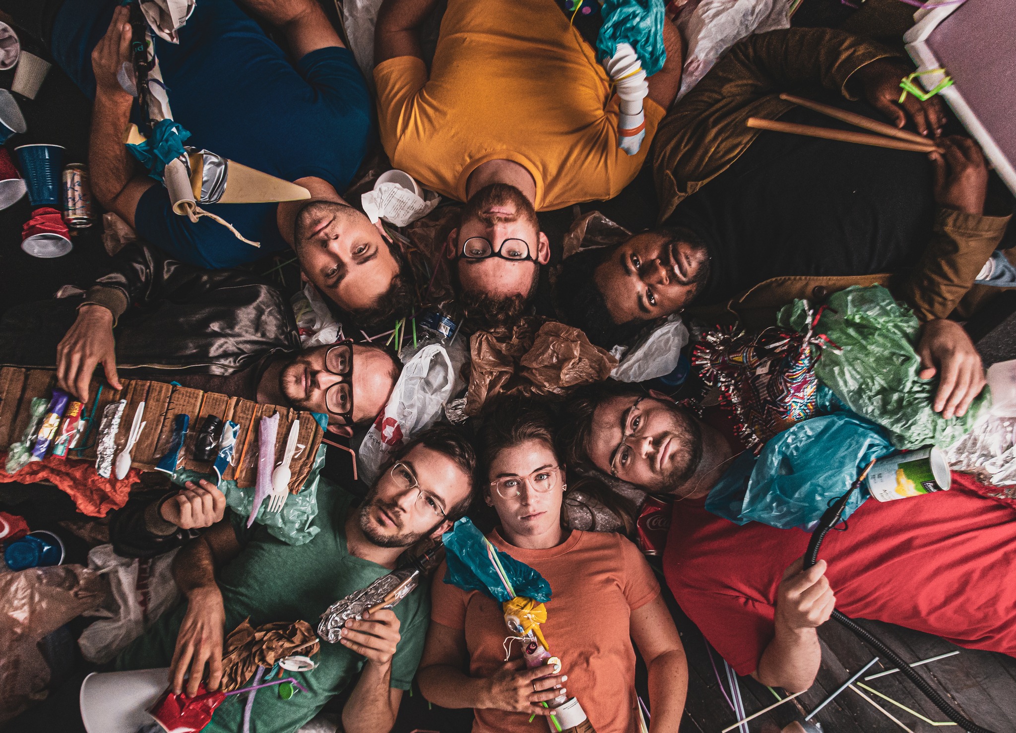 Seven people lay face-up in a circle, staring up at the camera with the image taken from above. They are surrounded by various miscellaneous objects, including straws, paper, plastic silverware, and empty cans.