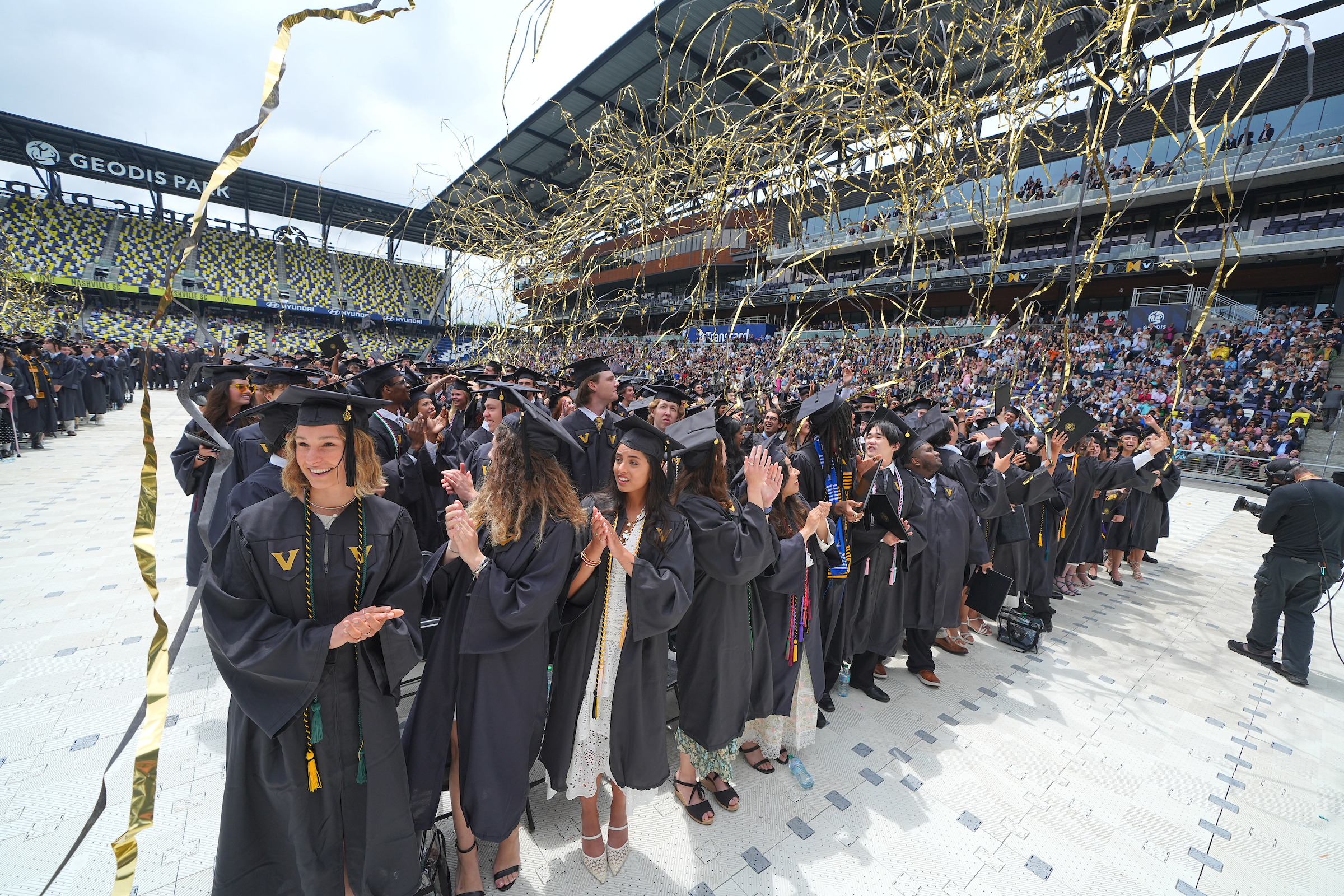 Black and gold streamers fly above a crowd of graduates standing in a stadium