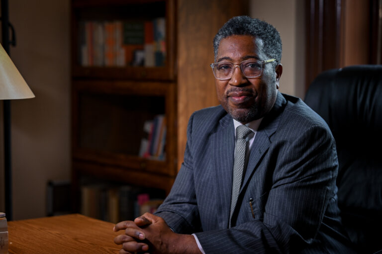 A man wearing a gray suit and glasses sits in a dimly lit office space with a book shelf in the background.