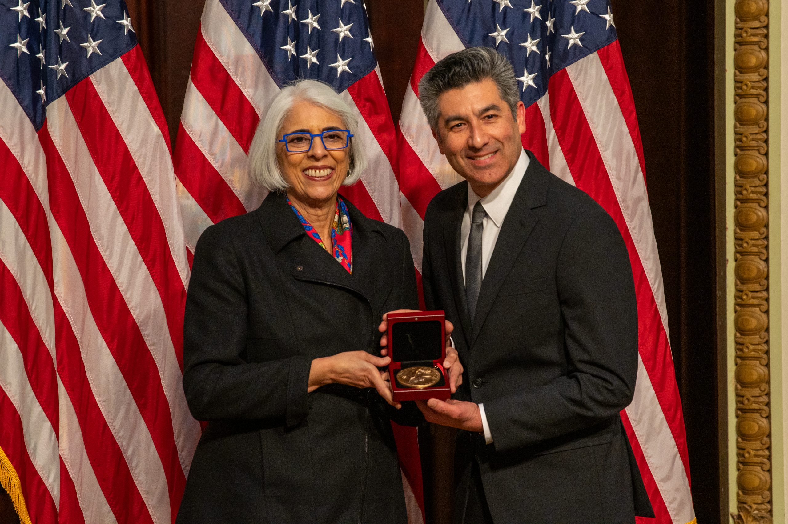 Keivan Stassun holds a medal with Arati Prabhakar and three U.S. flags as the backdrop