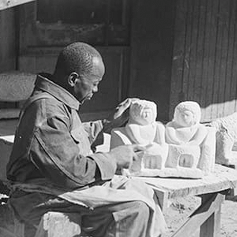Artist William Edmondson carving sculptures of human figures while seated at a workbench outdoors.