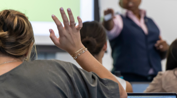 Pictured: Professor Jen Riley encourages student participation in her classes. This image shows a student is raising their hand in Riley's class. Riley is pointing toward the student to indicate it is their turn to speak.