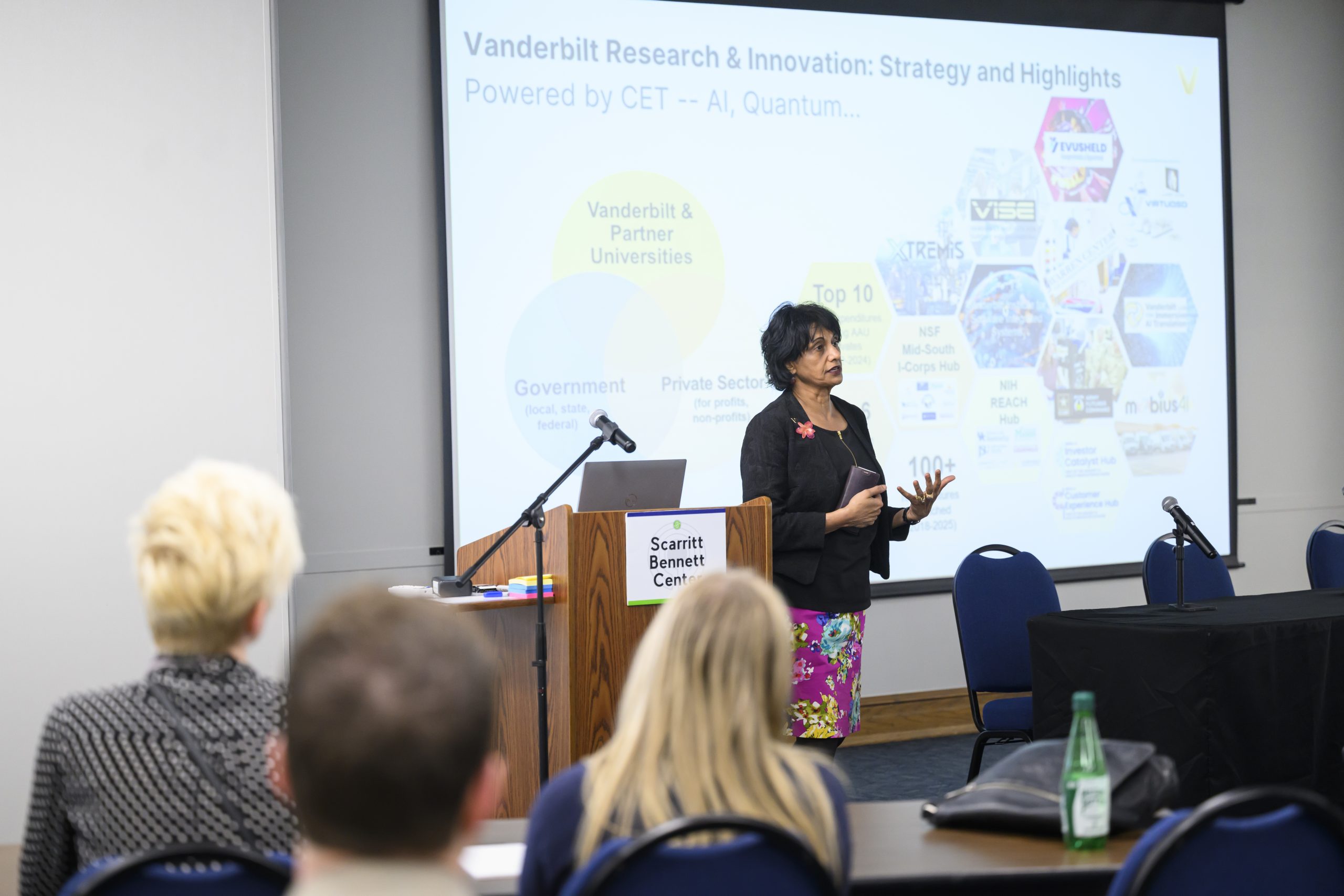 A woman speaking to a seated classroom with a presentation in the background