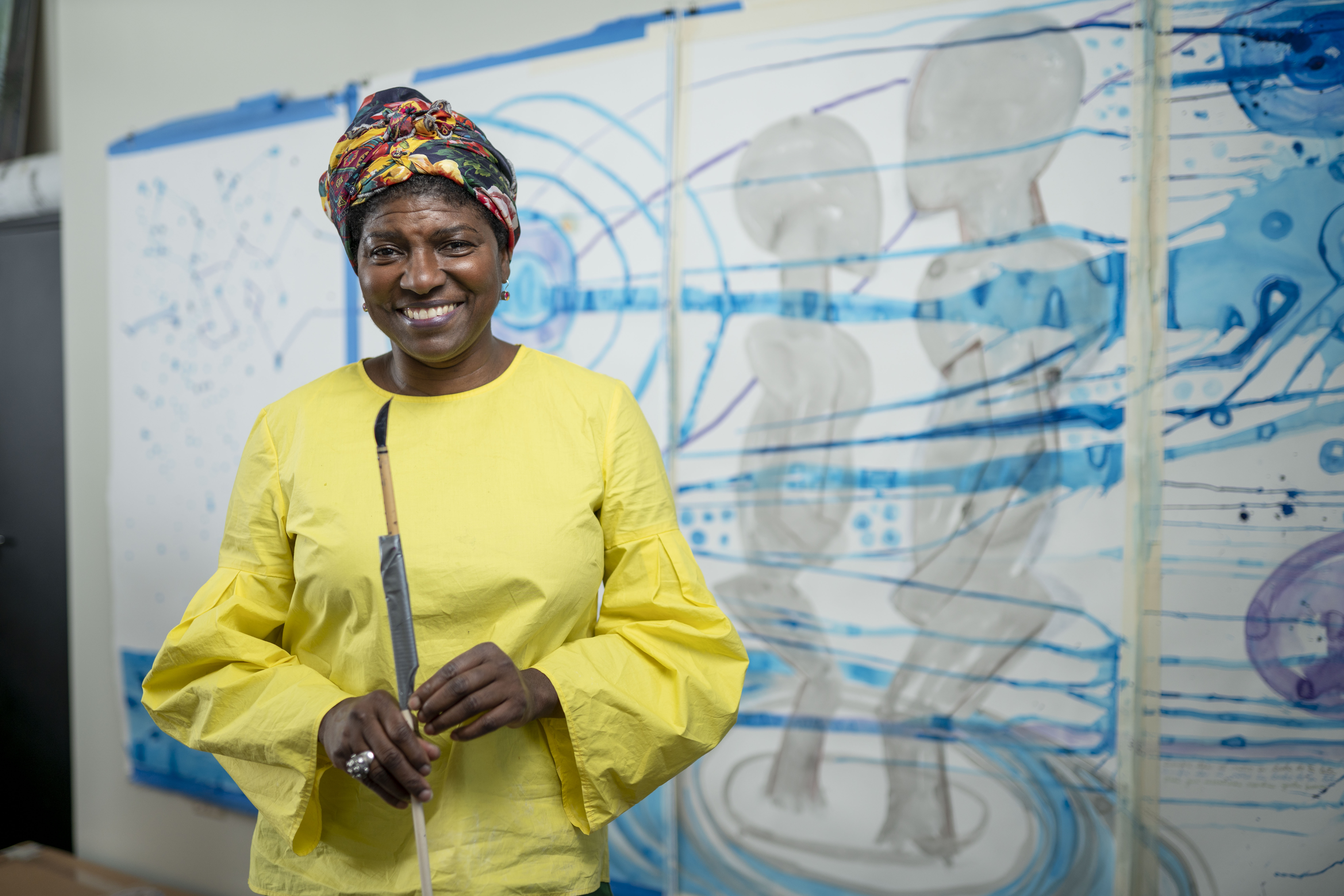 María Magdalena Campos-Pons smiles while holding a paintbrush in front of some of her artwork.