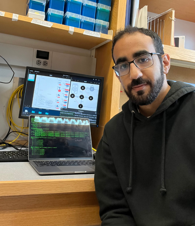 A man with short dark hair, a beard, and glasses sits at a lab workstation. He wears a black hoodie and looks toward the camera with a slight smile. Behind him are two computer screens—one showing a terminal with lines of code and the other displaying a flowchart with scientific diagrams. Shelves above the desk hold plastic lab containers labeled “200 uL Filter” and “1000 uL Filter.”