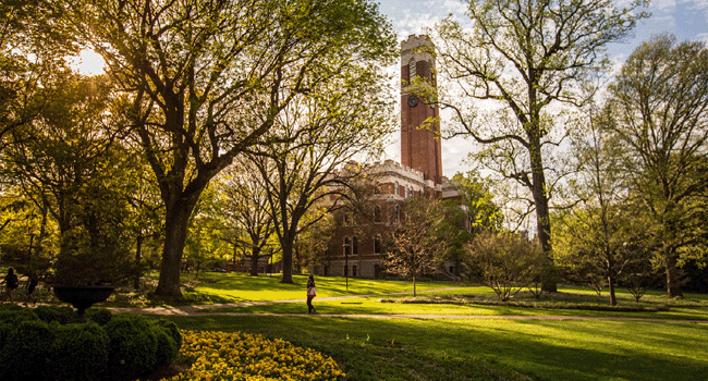 View of Vanderbilt's main campus with sun shining through trees and onto the grass with the bell tower of Kirkland Hall looming in the background as a student walks on a sidewalk path