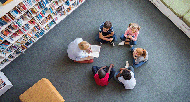 Children sit in a circle listening to an adult read to them in a peaceful library