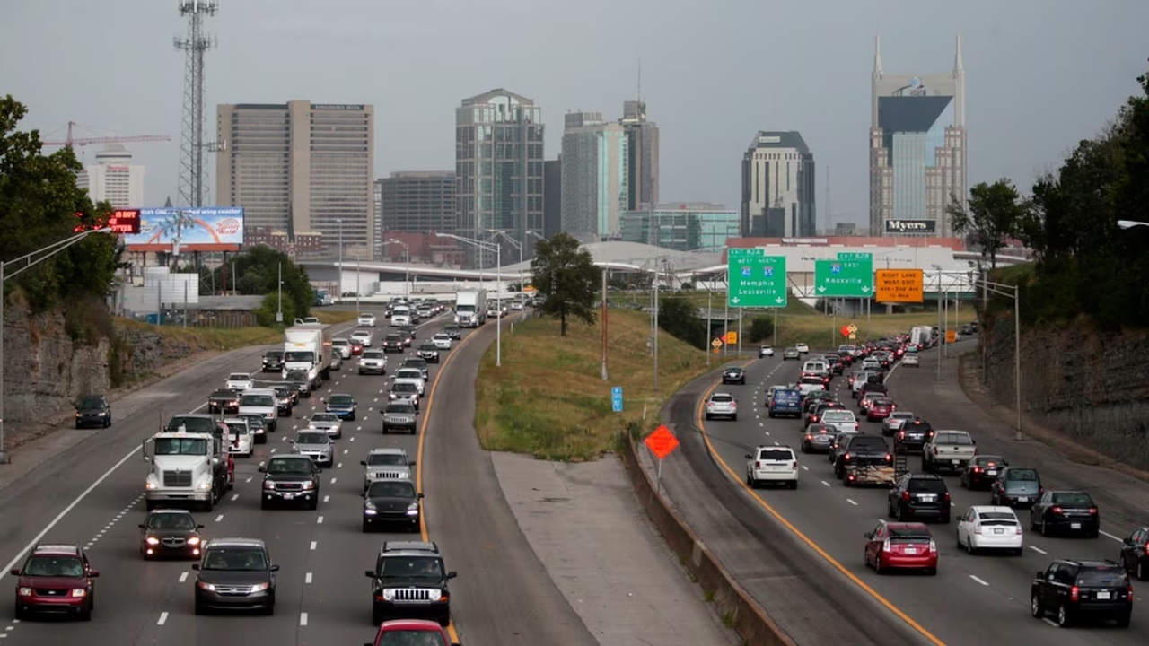 View of a busy highway in Nashville, Tennessee, with multiple lanes of traffic in both directions. The downtown skyline is visible in the background, featuring prominent buildings such as the AT&T 'Batman' Building. Road signs point to destinations like Memphis, Louisville, and Knoxville.