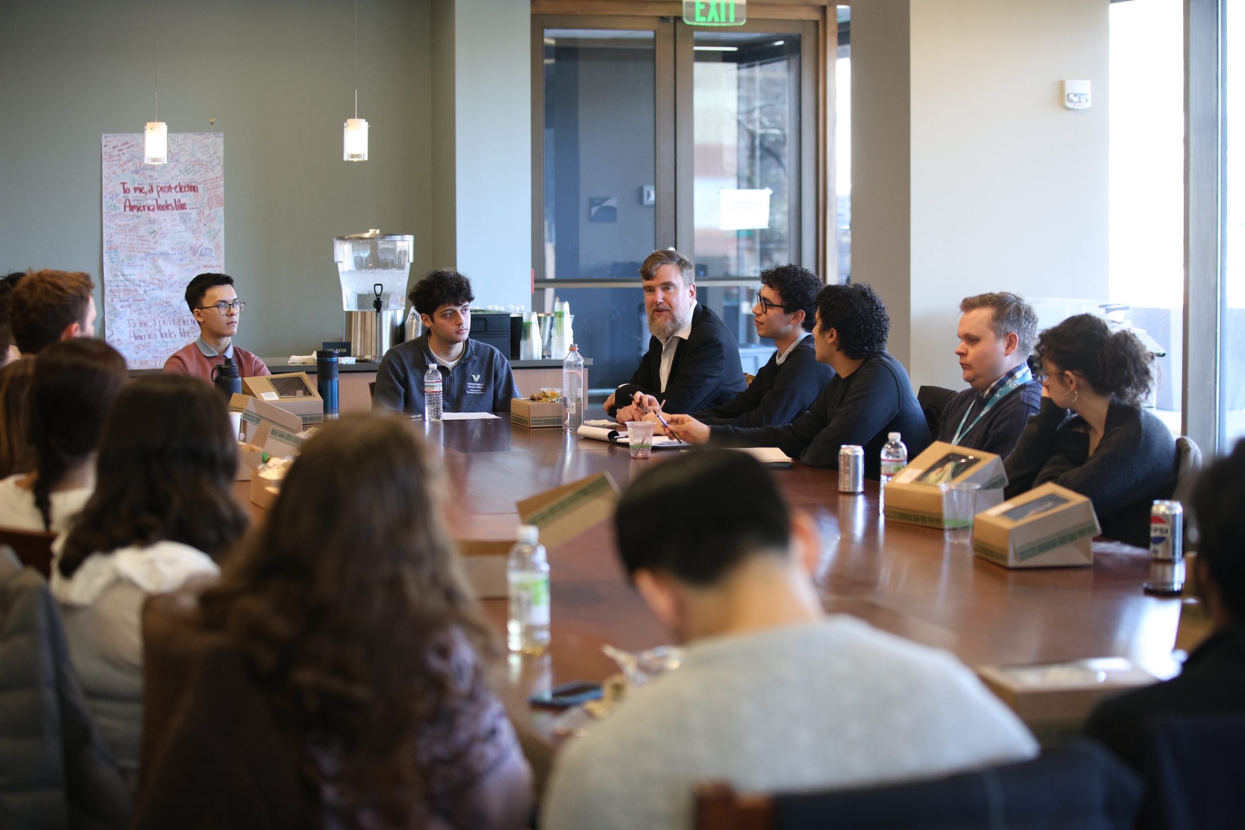 Students seated around a wooden table listening a man speaking. 