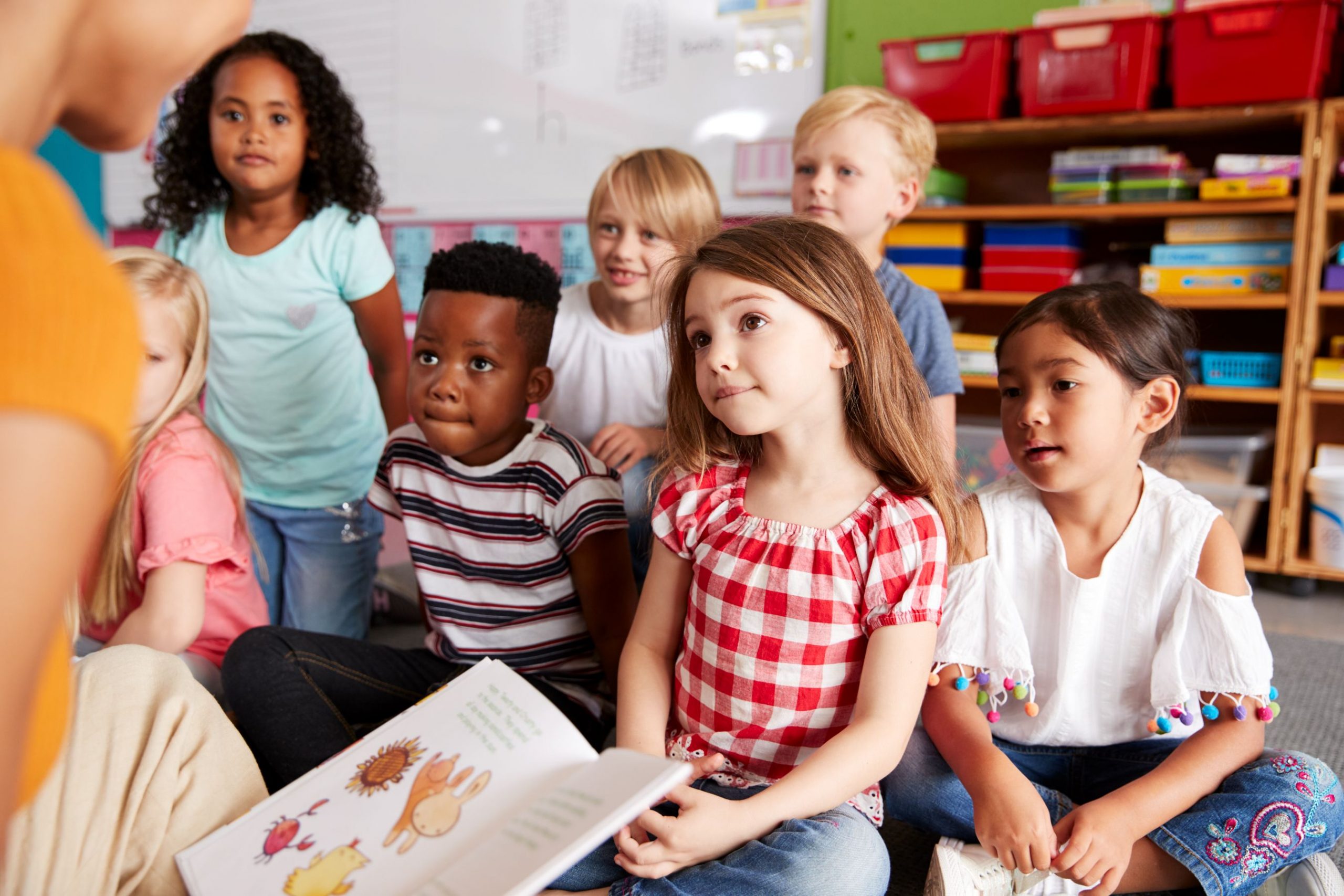 A group of young students sit on the floor and listen intently as a teacher reads a book to them.