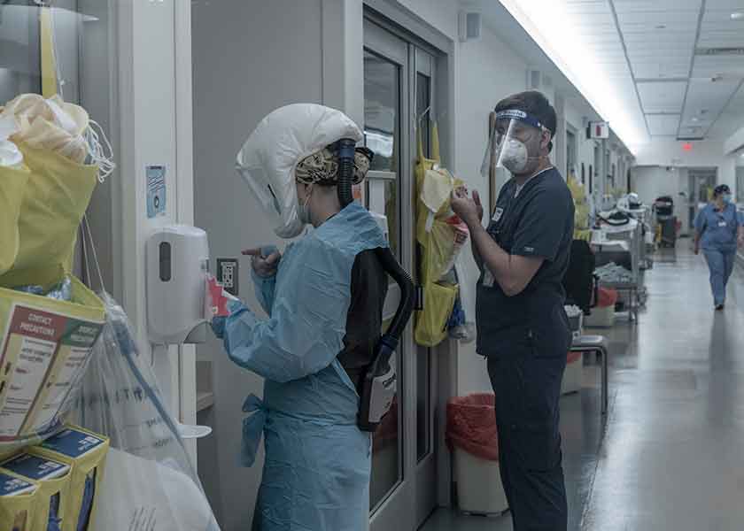 Nurse practitioner in protective gown and equipment prepares to enter a patient's room. A second health care professional is behind her, preparing to don his PPE.