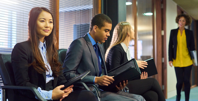 three interview candidates wait nervously in the reception of an office building. a member of the interview panel can be seen walking towards them.