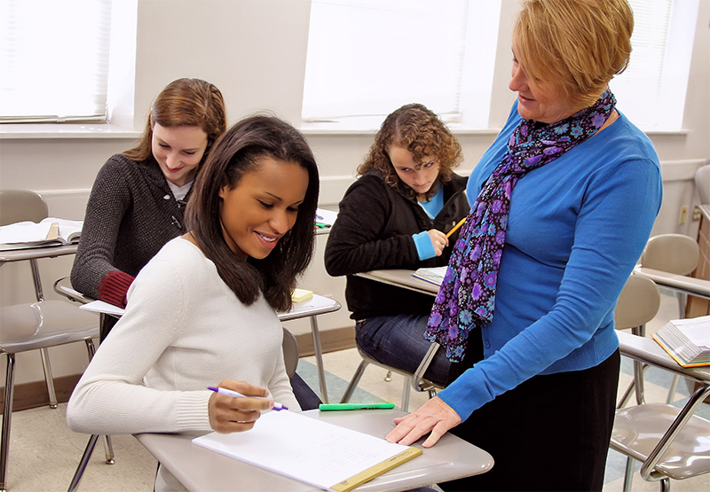 Students working at their desks and teacher pointing to Kyra's desk