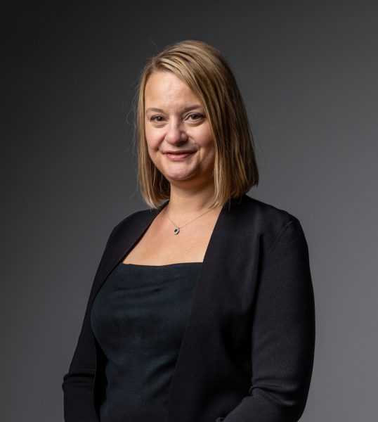 A woman in a black dress and jacket standing against a gray background