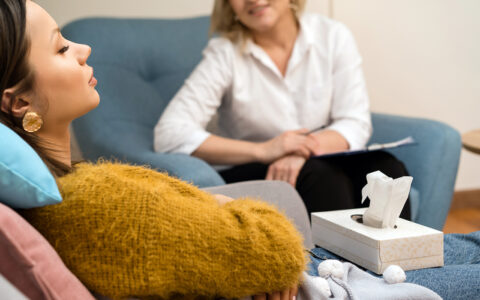 Young woman with closed eyes reclines on couch. In the background, out of focus, another woman is sitting in an armchair and observing