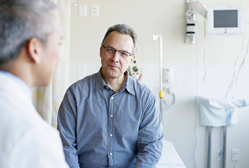 A middle-aged man faces the camera during a doctor's appointment, with the doctor's back to the camera.