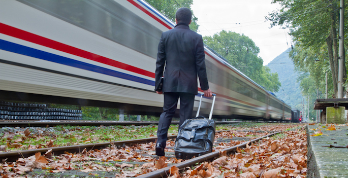 traveler dragging suitcase along train tracks as a missed train rushes past