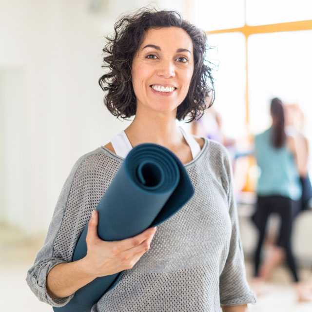Woman stands in an exercise studio holding a yoga mat.