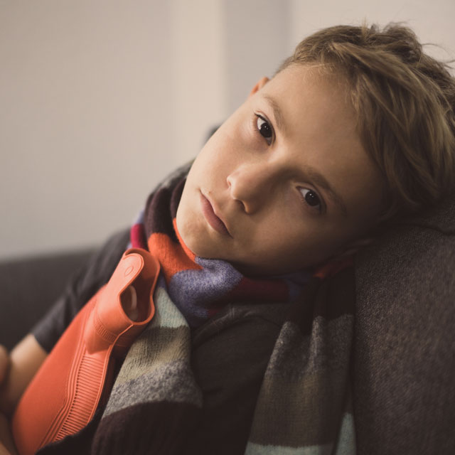A school-aged boy sits on a couch with a hot water bottle pressed to his stomach