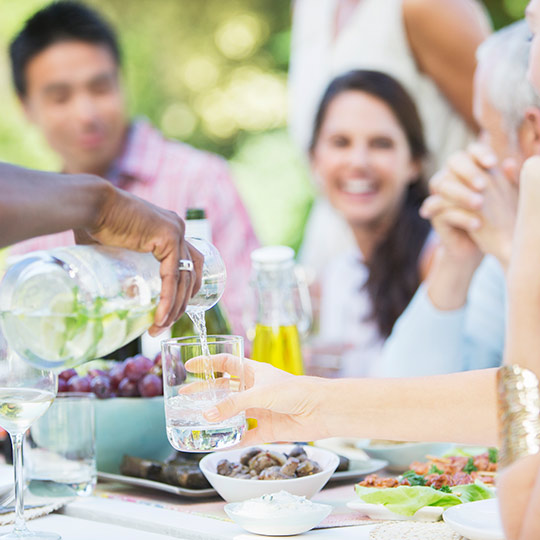 Group of people sitting around a table of food. A man in foreground fills a woman's water glass.
