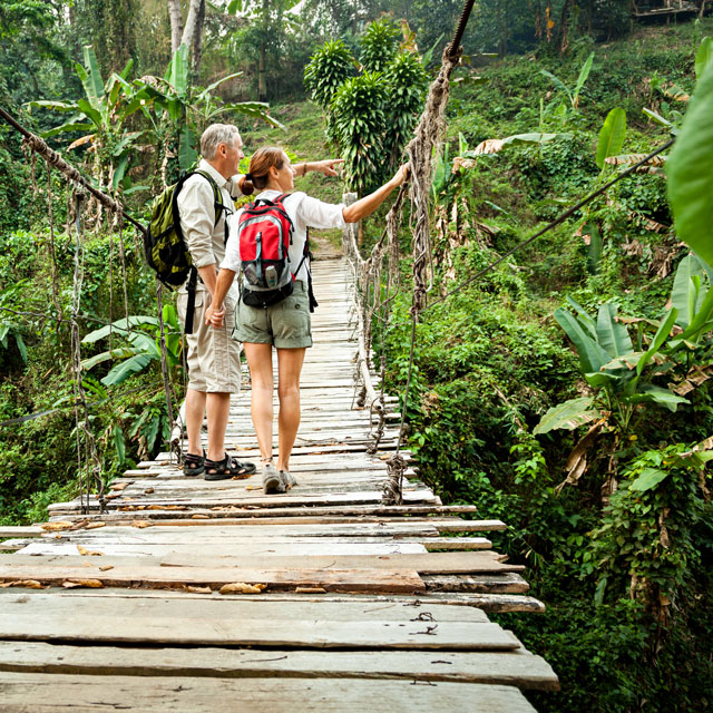 White middle aged couple stand on a plank bridge in a jungle.