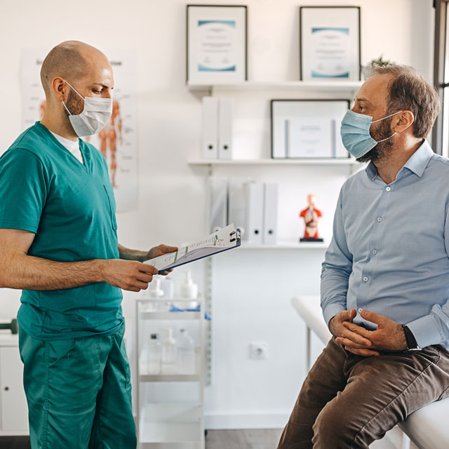 A male doctor in scrubs consults with a male patient in a clinic room, both wearing masks.
