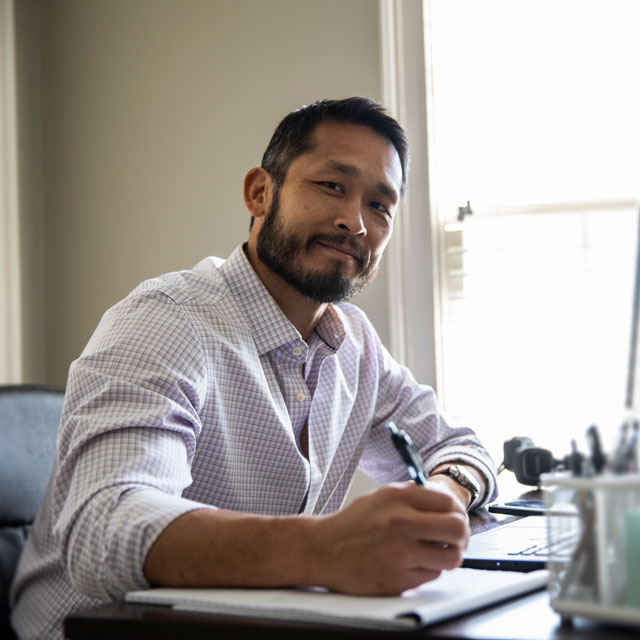 A middle-aged Asian man sits at a desk working in front of a laptop.