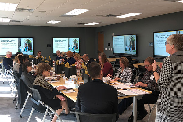 A group of people sit on four sides of a long table and listen to Senior Associate Dean Mavis Schorn talk. The room is modern and has large digital displays on all the walls.