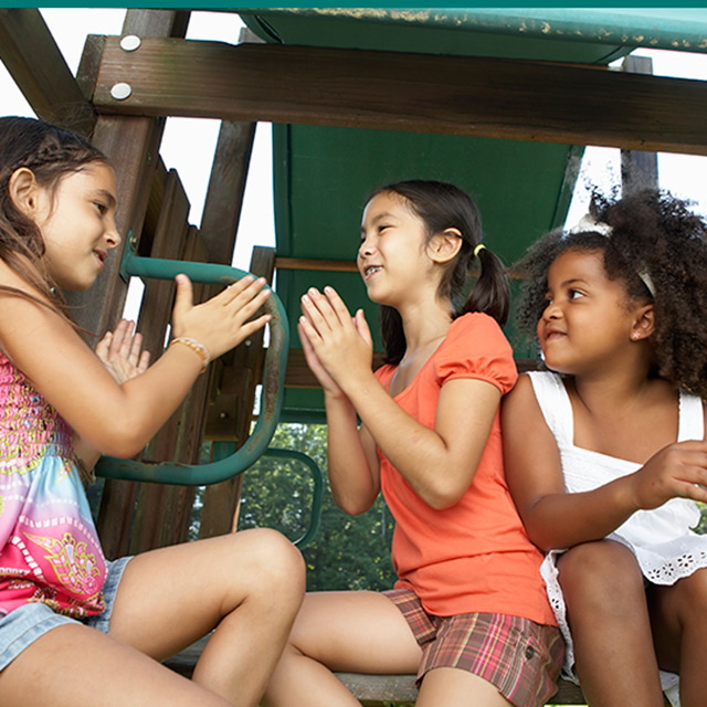 Three girls play patty cake sitting on a piece of playground equipment
