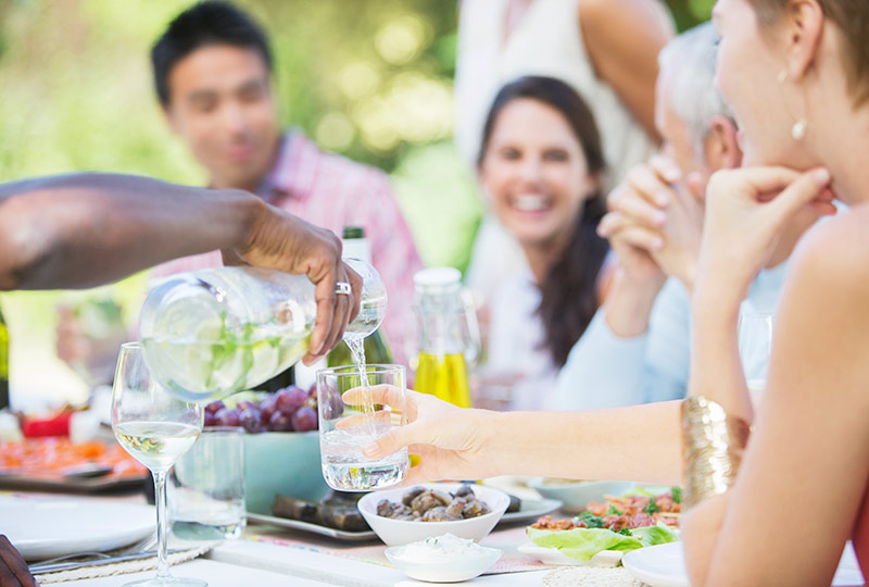 Group of people sitting around a table of food. A man in foreground fills a woman's water glass.