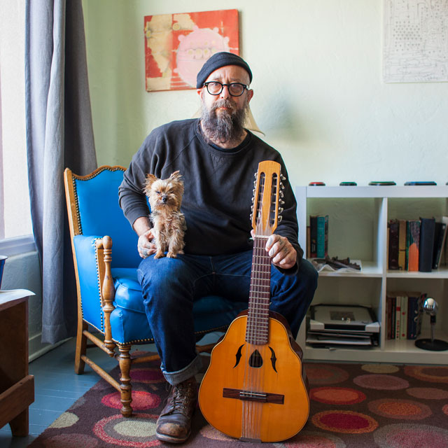 Older man sits holding a Yorkshire terrier on his lap and a mandolin at his feet.