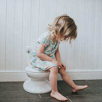 Toddler girl sits, fully clothed, on the closed lid of a child's potty seat.