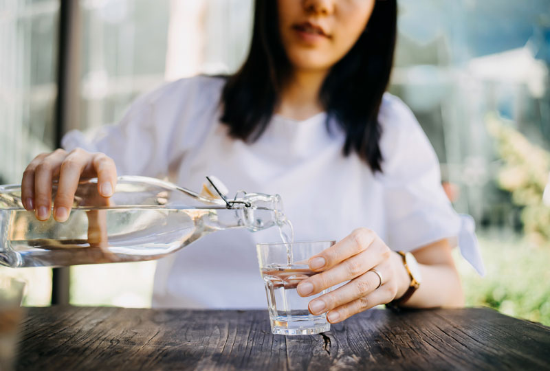 A woman pours water from a large bottle into a small drinking cup at an outdoor table.