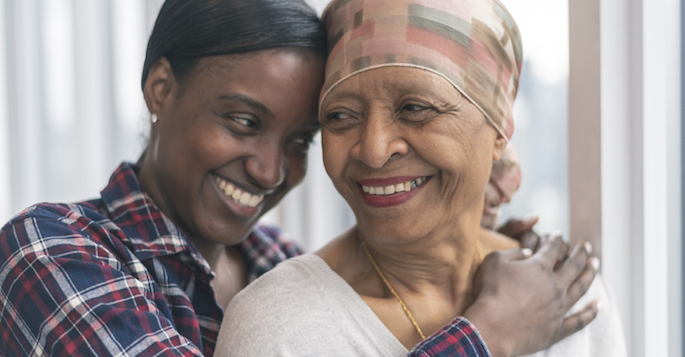 A black senior woman with cancer is wearing a scarf on her head. Her adult daughter is giving her a hug. Both women are smiling with gratitude and hope for recovery.