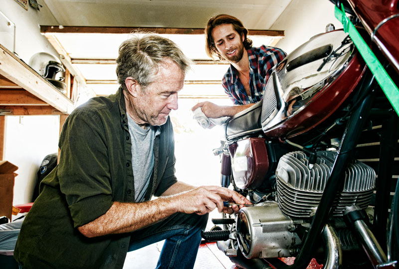 An older man kneels to work on a motorcycle engine while a younger man watches in a garage.