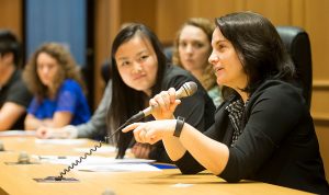 (Right) Prof. Tara McKay hold microphone, multiple students in the background focusing Prof. McKay speaking.