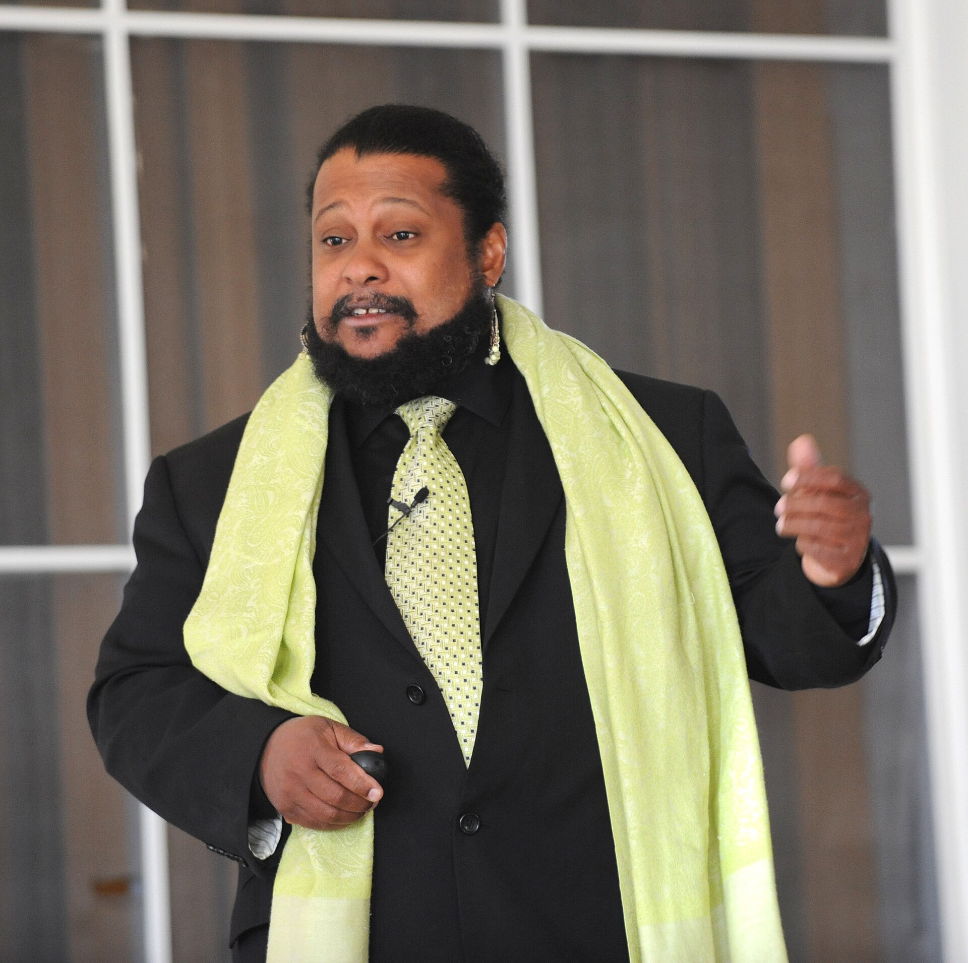 Tyrone Hayes speaks while gesturing with one hand during a presentation. He wears a black suit with a patterned green tie and a matching green scarf draped over his shoulders. A microphone is clipped to his tie, and a large earring is visible in his left ear. He stands in front of a window with dark curtains.