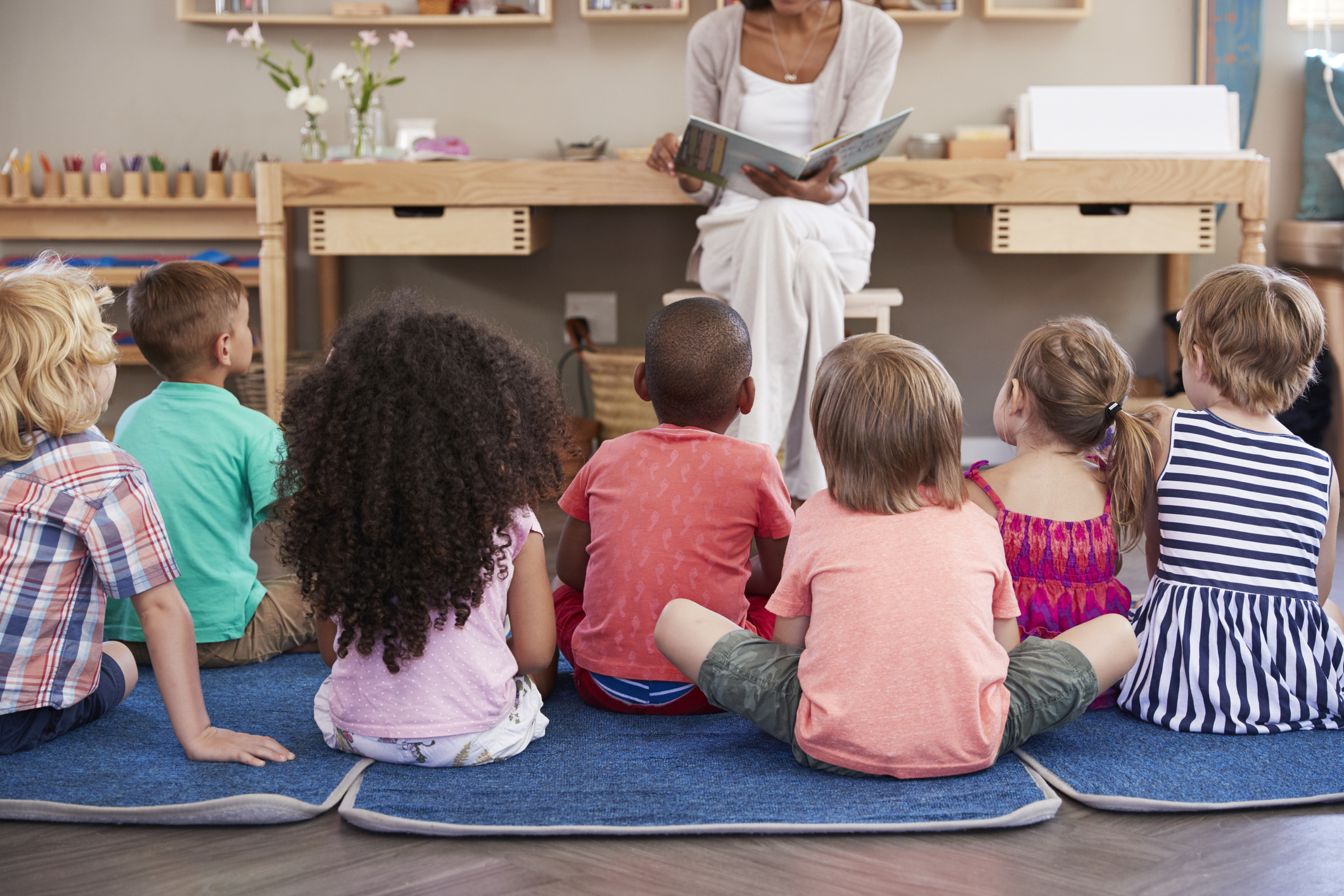Elementary school students sitting on the floor listening to their teacher read a book to them. 