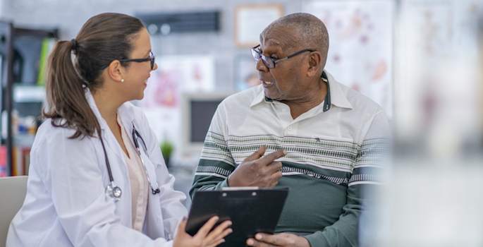 Older African American man and a young white female doctor discussing his health record in a doctor's office.