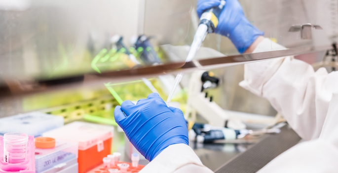 Close up photo of white-coated, gloved hands extracting fluid from a vial underneath a fume hood in a laboratory (Getty)