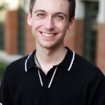 A man in a black and white shirt stands outside and smiles