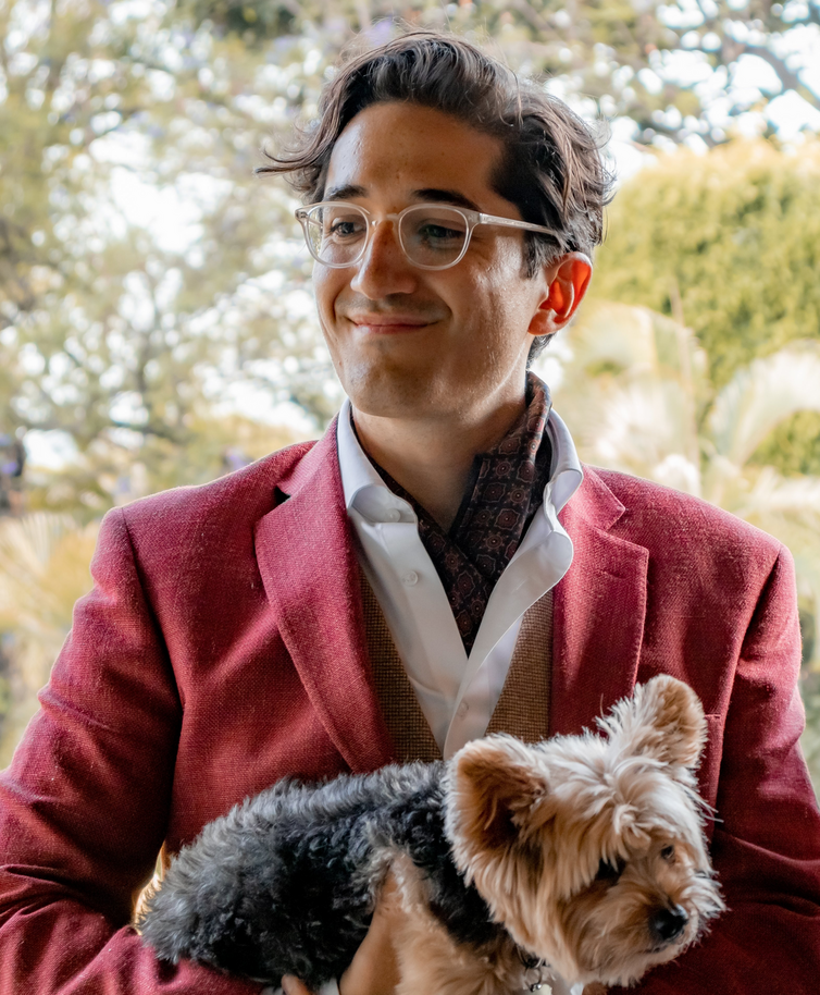 Carlos Oliver, smiling and looking off camera, holding a yorkshire terrier in his arms.