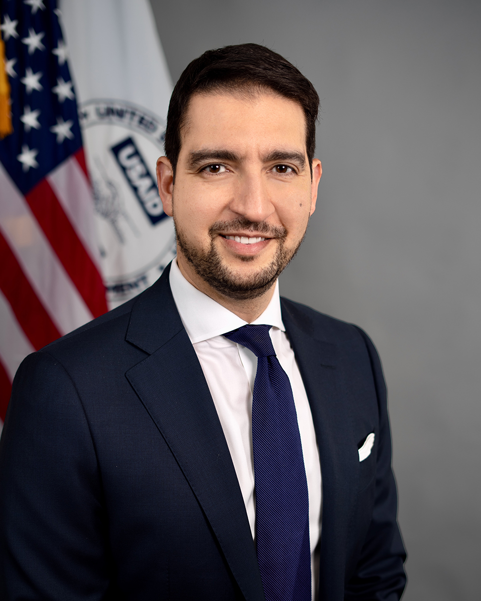 A young man in a suit standing in front of the U.S. flag and the USAID flag